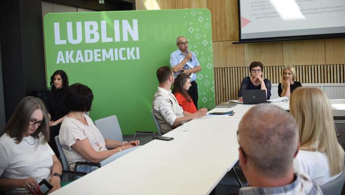 Pokaż zdjęcie: Uczestnicy spotkania w Lublinie siedzą przy stole konferencyjnym, a prelegent z mikrofonem prowadzi wystąpienie na tle zielonej ścianki z napisem „Lublin Akademicki”.