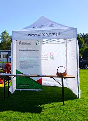 Zdjęcie przedstawia biały namiot promocyjny z napisem PFRON i naszym logo. Pod namiotem stoi roll-up naszego Centrum Informacyjnego dla Osób z Niepełnosprawnościami. Przed nim stoi stolik, na którym leżą ulotki informacyjne i stoi koszyk.