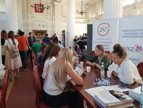 Duża sala, na niej targi pracy, część osób siedzi za stołami, pozostali chodzą, widoczne banery instytucji i pracodawców
