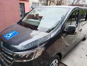 Pokaż zdjęcie: Na parkingu stoi zaparkowany samochód. Naklejona jest na nim naklejka, która informuje, że pojazdem są przewożone osoby na wózkach inwalidzkich. 