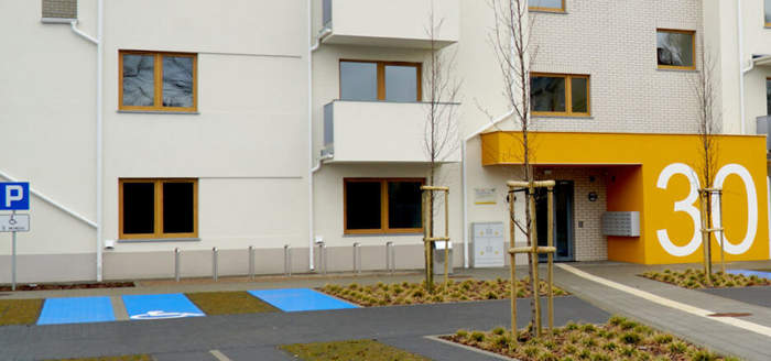 Pokaż zdjęcie: Kilkupiętrowy blok, przed nim dwa miejsca oznaczone dla niepełnosprawnych. Wejście w jaskrawym kolorze z dużym napisem 30.