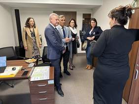 Pokaż zdjęcie: W biurze dwóch mężczyzn w marynarkach i cztery kobiety. Jedna z kobiet stoi tyłem przy szafie i tłumaczy coś pozostałym ludziom.