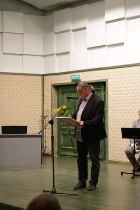 Pokaż zdjęcie: Na scenie stoi mężczyzną w ciemnym garniturze i jasna koszulą wyciągniętą ze spodni, włosy ma zaczesane w kucyk, na nosie ma okulary. Trzyma dyplom wraz z medalem oraz bukiet kwiatów.