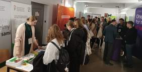 Pokaż zdjęcie: Grupa studentów na korytarzu uczelni, gdzie swoje stoiska mają wystawcy Targów Pracy. Z przodu stoisko Oddziału Wielkopolskiego PFRON, przy którym kobieta udziela informacji uczestnikowi Targów. Na stoliku wyłożone są materiały informacyjne.