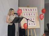 Kobieta przemawia przy mównicy podczas gali „Otwarte Serca Roku 2026”. W tle tablica z nazwą wydarzenia, czerwone balony i statuetki przygotowane dla wyróżnionych. Sala udekorowana w biało-czerwonych barwach.