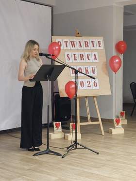 Kobieta przemawia przy mównicy podczas gali „Otwarte Serca Roku 2026”. W tle tablica z nazwą wydarzenia, czerwone balony i statuetki przygotowane dla wyróżnionych. Sala udekorowana w biało-czerwonych barwach.