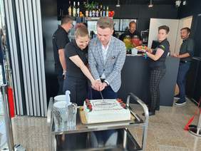 Pokaż zdjęcie: Wewnątrz kawiarni mężczyzna z kobietą wspólnie trzymają nóż i kroją tort z nazwą kawiarnia Alternatywa 21 obok stoją talerzyki i sztućce