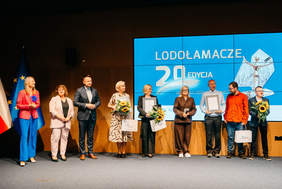Pokaż zdjęcie: Grupa dziewięciu dorosłych osób stoi na scenie przed dużym, jasnoniebieskim ekranem z napisem "LODOŁAMACZE 20 EDYCJA". Jest to prawdopodobnie scena ceremonii wręczenia nagród. Ludzie stoją w rzędzie, ubrani w stroje galowe lub biznesowe, a kilka z nich trzyma dyplomy, nagrody (prawdopodobnie statuetki) lub bukiety słoneczników. Po lewej stronie widoczne są fragmenty flag, w tym flagi Polski i Unii Europejskiej. Ostatnia osoba po prawej trzyma również małą, żółtą maskotkę