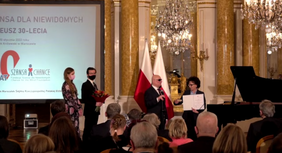 Pokaż zdjęcie: Na scenie Marszałek Sejmu RP Pani Elżbieta Witek odbiera IDOLA SPECJALNEGO od Pana Marka Kalbarczyka. W tle telebim z logotypem fundacji. W dole zdjęcia, siedząca publiczność.