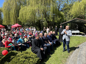 Pokaż zdjęcie: Na polanie siedzi dużo osób na ławkach w pierwszym rzędzie biskup wójt i inni zaproszenie goście. naprzeciw nich stoi mężczyzna ubrany w jasną marynarkę ciemne spodnie trzyma mikrofon i przemawia do nich. dookoła wysokie drzewa jest piękna sł