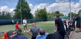 Pokaż zdjęcie: Trawiasty kort tenisowy. Kilkanaście osób na wózkach inwalidzkich ogląda pokaz gry w tenisa na wózku.