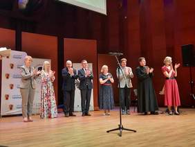 Pokaż zdjęcie: Na scenie stoi osiem osób: cztery kobiety i czterech mężczyzn. Biją brawo i uśmiechają się. Przed nimi stoi mikrofon. 