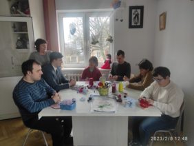 Pokaż zdjęcie: Trzy kobiety i trzech mężczyzn siedzi przy stole i zajmują się robótkami ręcznymi. Jedna kobieta stoi.