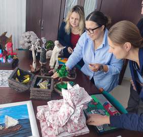 Pokaż zdjęcie: Trzy kobiety pochylają się nad stołem na którym wyeksponowane są rzeźby.