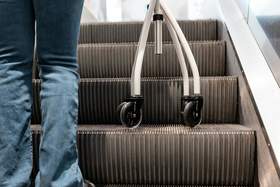 Na ruchomych schodach stoi człowiek, któremu widać tylko tył nóg. Obok jest laska inwalidzka trójnóg. Osoba jest ubrana w niebieskie dżinsy. 