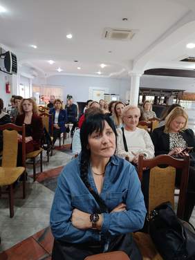 Pokaż zdjęcie: Na sali z kolumnami siedzą ludzie na krzesłach. W pierwszym rzędzie siedzi kobieta w niebieskiej bluzce i uważnie słucha.