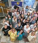 Grupa około dwudziestu pięciu uśmiechniętych osób, głównie kobiet, sfotografowana z góry w jasnym wnętrzu. Stoją blisko siebie, patrzą w obiektyw i machają rękami w stronę aparatu. W tle widoczny jest granatowy baner z żółtym sercem i białym napisem „Fundacja Pokonać Endometriozę”.