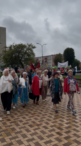 Pokaż zdjęcie: Grupa kobiet i mężczyzn idzie chodnikiem podczas wydarzenia. Uczestnicy są ubrani w kolorowe stroje, część osób trzyma flagi i transparenty. W tle znajdują się drzewa, budynki mieszkalne i zachmurzone niebo.