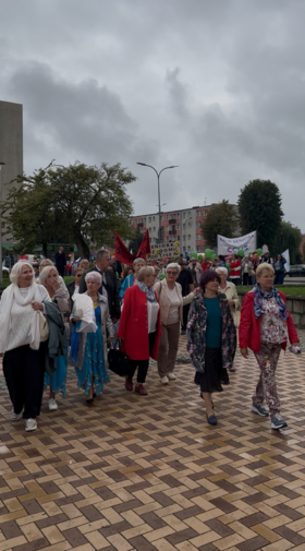 Grupa kobiet i mężczyzn idzie chodnikiem podczas wydarzenia. Uczestnicy są ubrani w kolorowe stroje, część osób trzyma flagi i transparenty. W tle znajdują się drzewa, budynki mieszkalne i zachmurzone niebo.
