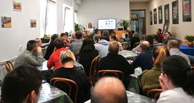 Pokaż zdjęcie: Sala wypełniona dorosłymi uczestnikami siedzącymi przy stołach, zwróconymi w stronę prowadzącego. Prelegent stoi z przodu z mikrofonem w dłoni, obok ekranu z wyświetloną prezentacją. W tle widać rośliny doniczkowe, drzwi oraz ściany ozdobione oprawionymi fotografiami.