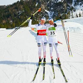 Dwóch narciarzy na trasie biegów w uścisku, na tle gór, każdy z uniesioną jedną ręką, uśmiechają się, słoneczny dzień