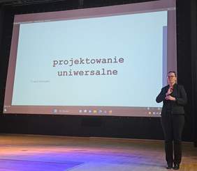 Po lewej stronie stoi Pani tłumaczka języka migowego, a w tle widoczny slajd z wykładu dr. Jakuba Elwertowskiego pt. "Projektowanie uniwersalne"