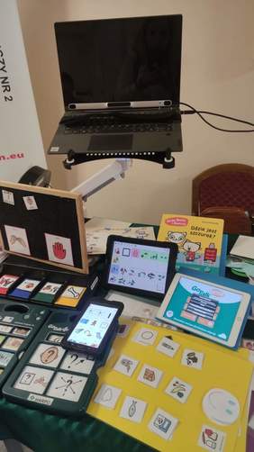Pokaż zdjęcie: Na stole leżą różne urządzenia takie jak laptopy, tablety, urządzenia te wspierają mowę oraz piktogramy ułatwiające komunikację