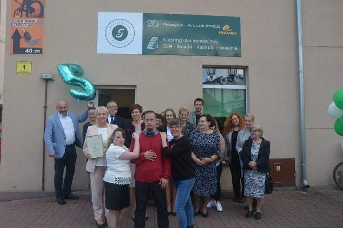 Pokaż zdjęcie: Grupa kobiet i mężczyzn stoi przed budynkiem Spółdzielni Socjalnej „Na piątkę”. Jeden z mężczyzn trzyma balon w kształcie cyfry 5. Na ścianie budynku zawieszony jest baner informujący o charakterze działalności spółdzielni.