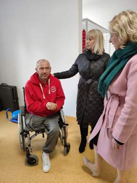 Pokaż zdjęcie: Zdjęcie w sali rehabilitacji, dwie kobiety w wierzchnich ubraniach rozmawiają z mężczyzną siedzącym na wózku inwalidzkim.