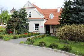 Pokaż zdjęcie: Zdjęcie od frontu jednopiętrowego budynku w kolorze białym z czerwonym dachem, siedziby środowiskowego domu samopomocy, w otoczeniu zieleni