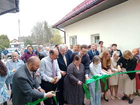 Przed domem mieszkalnym, w którym mają zamieszkać osoby z niepełnosprawnościami, stoją ludzie i uroczyście przecinają wstęgę. Wszyscy są elegancko ubrani i w dobrych humorach. 