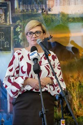 Pokaż zdjęcie: Kobieta stojąca przed dwoma mikrofonami wygłasza oficjalne przemówienie podczas wydarzenia publicznego. Ubrana jest w elegancką bluzkę. Za nią znajduje się kolorowa plansza przedstawiająca regionalne krajobrazy. 