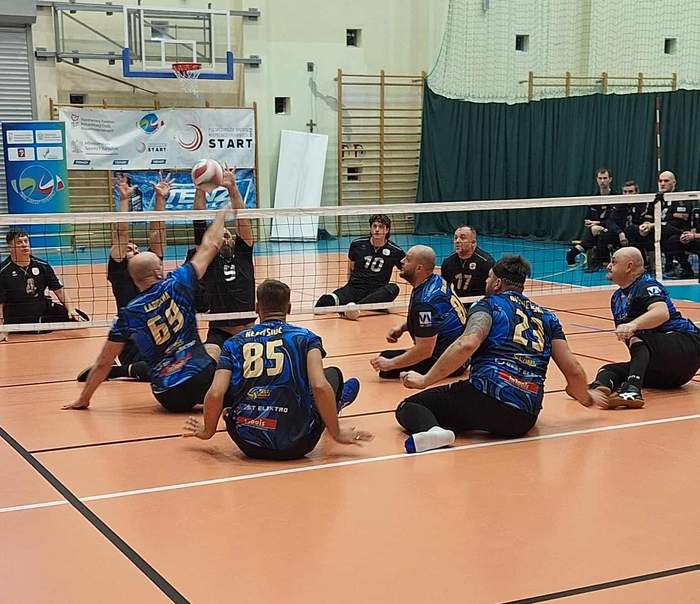 Pokaż zdjęcie: Dynamiczna scena z meczu Piłki Siatkowej na Siedząco. Kilku zawodników w czarno-niebieskich strojach, siedząc na podłodze boiska, bierze udział w akcji przy siatce. Jeden z graczy w powietrzu atakuje piłkę, a inni w pozycji siedzącej są gotowi do obrony lub przyjęcia. Wszyscy zawodnicy mają koszulki z numerami, a jeden z nich (numer 85) ma napis "READY SWC". Na drugim planie, za siatką, siedzi drużyna przeciwna w czarnych strojach. Na ścianach w tle widoczne są banery promujące wydarzenie, w tym logo START i informacja o Polskim Związku Sportu Niepełnosprawnych.