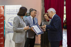 Pokaż zdjęcie: Mężczyzna w garniturze wręcza certyfikat oprawiony w aluminiową ramę. Naprzeciw stoi siwy mężczyzna w granatowym garniturze i okularach. W ręku trzyma statuetkę. Obok stoi kobieta trzymająca różę.