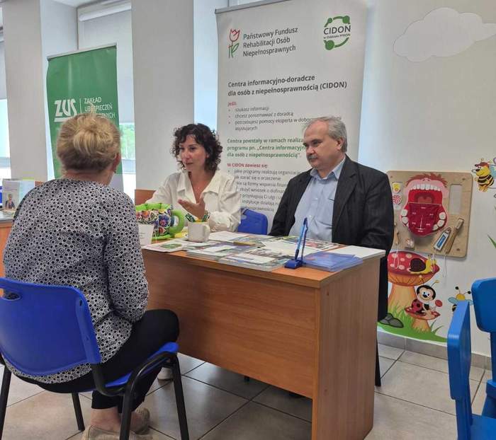 Pokaż zdjęcie: Przy biurku w sali NFZ siedzą trzy osoby, które rozmawiają. Na stole leżą ulotki, w tle rollup z logo CIDON i PFRON.
