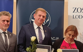 Widzimy popiersia trzech uśmiechniętych osób, od lewej szpakowaty mężczyzna, w środku wysoki o włosach blond, trzyma białą różę i lekko przechyla głowę w bok, obok kobieta z pochyloną głową i opuszczonym wzrokiem jakby czytała