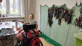 Pokaż zdjęcie: Na ścienie pracowni rozwieszona siatka maskująca. Przy stoliku siedzi kobieta, która wykonuje kolejne elementy siatki