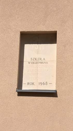 Pokaż zdjęcie: Fragment elewacji budynku z tablicą pamiątkową zawierającą napis informujący o roku powstania szkoły.