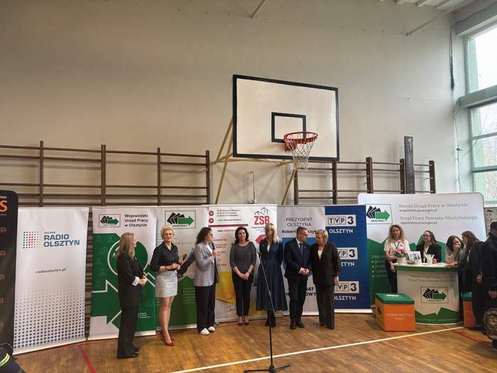 Pokaż zdjęcie: Siedem osób stoi na środku Sali przy mikrofonie na tle banerów instytucji w sali gimnastycznej; ubrani w formalne stroje; jedna z osób przemawia, pozostali słuchają.