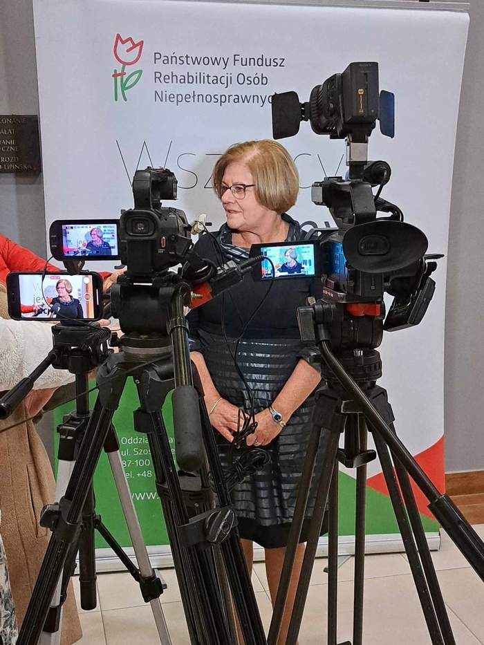 Dyrektor Kujawsko-Pomorskiego Oddziału PFRON Aleksandra Gierej udziela wywiadu - przed nią stoją sttaywy z kamerami, a za nią baner PFRON-u