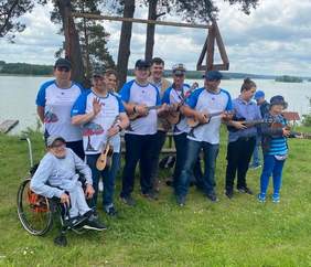 Pokaż zdjęcie: Kilkanaście uśmiechniętych osób ubranych w koszulki z napisem Warsztat Terapii Zajęciowej stoi na tle jeziora. 6 osób ma w rękach gitary