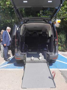 Pokaż zdjęcie: Na kopercie stoi samochód przystosowany do przewozu osoby poruszającej się na wózku inwalidzkim. Tylna klapa auta jest otwarta i widać miejsce na wózek inwalidzki. Po lewej stronie pojazdu stoją dwie osoby. 