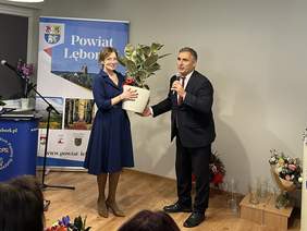 Pokaż zdjęcie: Kobiecie w eleganckiej sukience mężczyzna w ciemnym garniturze wręcza piękny kwiat w białej donicy.