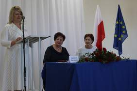 Pokaż zdjęcie: W sali siedzą dwie kobiety przy stoliku .Jedna kobieta stoi przed mikrofonem i przemawia. Za siedzącymi paniami stoją dwie flagi Polski i Unii Europejskiej 