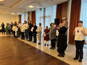 Pokaż zdjęcie: W sali bankietowej stoją w szeregu ludzie. Są to laureaci konkursu plastycznego "Sztuka Osób Niepełnosprawnych". Trzymają w dłoniach statuetki, dyplomy i nagrody. 