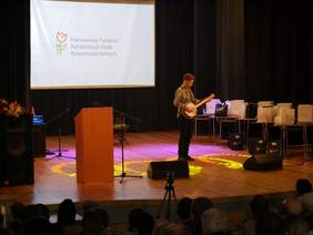 Pokaż zdjęcie: na scenie stoi młody mężczyzna i gra na instrumencie muzycznym podobnym do gitary obok stoją stoliki na których ustawione są nagrody w tle na białym ekranie wyświetlone jest logo PFRON