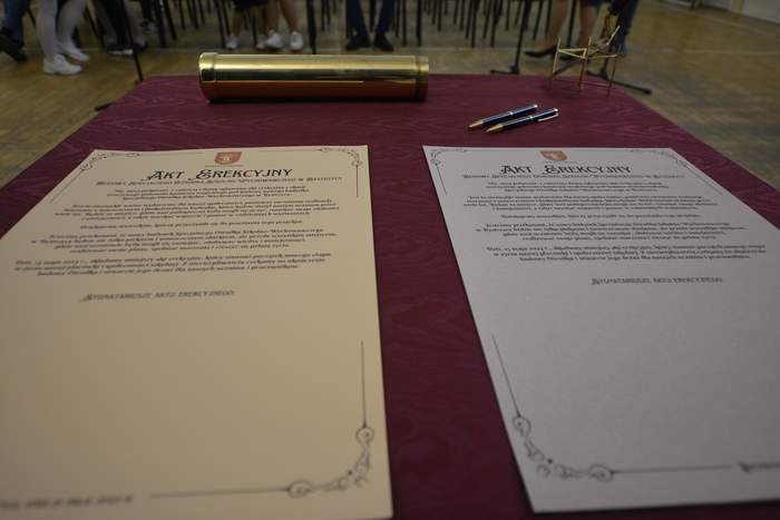Pokaż zdjęcie: Na stole przykrytym bordowym obrusem leża dwa dokumenty zatytułowane Akt Erekcyjny