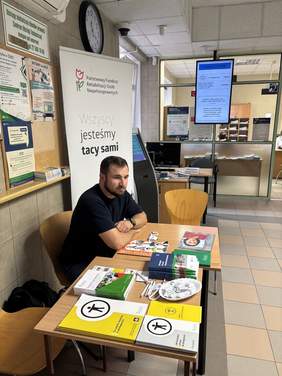 Pokaż zdjęcie: Mężczyzna siedzi przy stoliku z rozłożonymi broszurami, ulotkami i dokumentami. Za nim widoczny jest stojący baner oraz wnętrze budynku.
