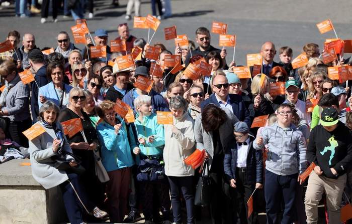 Pokaż zdjęcie: Duża grupa osób trzymających w ręku pomarańczowe chorągiewki, wśród zgromadzonych osób są osoby na wózkach inwalidzkich.