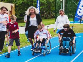 Pokaż zdjęcie: Dwóch chłopców na wózkach inwalidzkich ściga się po niebieskiej nawierzchni. Obok biegnie kobieta i chłopiec którzy kibicują. W tle flagi miasta Gdynia.
